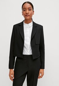 Schwarze, gekürzte Blazerjacke mit strukturierten Revers, offener Vorderseite und langen Ärmeln, über einem weißen Kragenhemd getragen. Glattes Material und taillierte Passform.