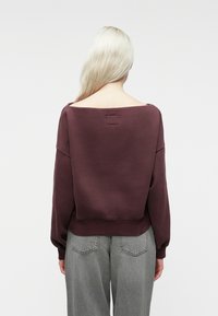 Šviesiaplaukis asmuo, vilkintis laisvą bordo spalvos megztinį be pečių ir pilkas aukštakulnės kelnes, matomas iš nugaros prieš paprastą foną.