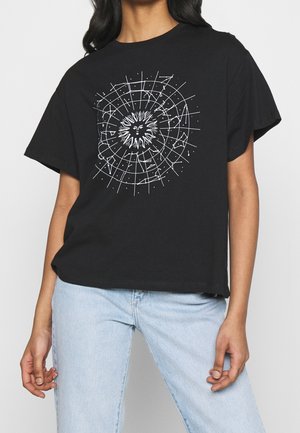 Person iført en sort t-shirt med et hvidt sol- og geometrisk gitterdesign, kombineret med lyseblå jeans, stående mod en ensfarvet baggrund.