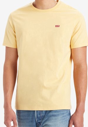 Homme portant un t-shirt uni jaune clair à manches courtes avec un petit logo rouge Levi's sur la poitrine gauche, associé à un jean bleu.