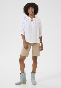 Witte linnen blouse met opgeblazen mouwen en een sleutelgat strikdetail, gecombineerd met beige shorts en blauwe cowboylaarzen met decoratieve stiksels.