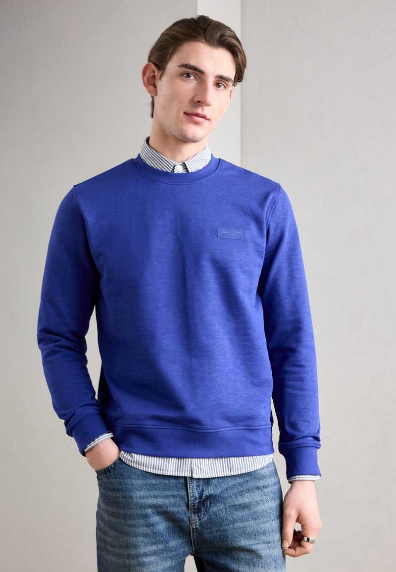 Felpa blu con polsini e orlo a costine, dotata di un'etichetta logo testurizzata. Indossata sopra una camicia a righe, abbinata a jeans blue.