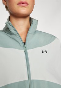 Chaqueta de color verde claro con cremallera y cuello alto, con un logo negro. La tela tiene una textura suave y secciones blancas contrastantes en el cuerpo.