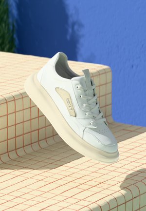 Witte leren sneaker met een crèmekleurige zool, geperforeerde neus en een subtiele beige accent. Beschikt over een logo aan de zijkant en platte veters.