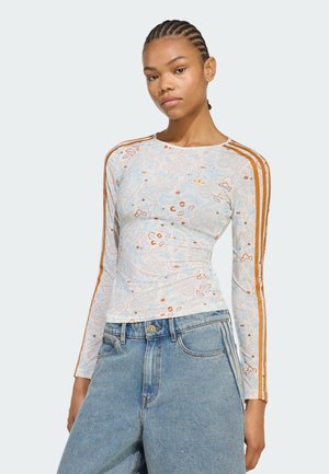 Mujer con el pelo trenzado que lleva un top blanco de manga larga con flores y rayas marrones en las mangas y vaqueros azul de talle alto sobre un fondo blanco.