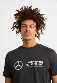 Czarna bawełniana koszulka z białym logo Mercedes-Benz i napisem "ZESPÓŁ FORMUŁY JEDNEJ AMG PETRONAS". Klasyczny krój z okrągłym dekoltem, krótkie rękawy.