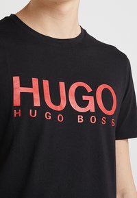 Schwarzes Baumwoll-T-Shirt mit auffälligem, rotem Drucktext, der "HUGO HUGO BOSS" über die Brust zeigt, mit Rundhalsausschnitt und kurzen Ärmeln.