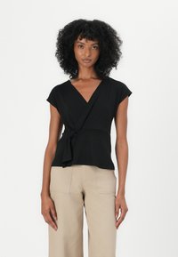 Top negru cu mâneci scurte, decolteu în V și un accent legat la talie. Purtați cu pantaloni largi bej. Textura materialului este netedă.