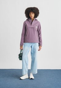 Casaco anorak roxo com capuz, bolso canguru, punhos elásticos, combinado com jeans azul claro e ténis brancos, segurando uma bolsa verde.