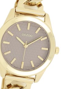 Montre en métal doré avec un cadran rond, une aiguille grise texturée, des index dorés et un bracelet à maillons. Nom de la marque "OOZOO" affiché.