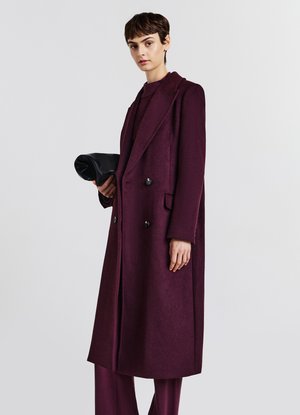 Manteau bordeaux à double boutonnage avec revers, poches latérales et détails en boutons noirs, assorti à un sac à main noir.