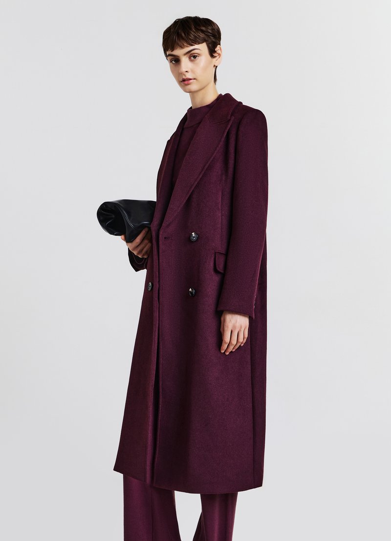 Cappotto bordeaux doppiopetto con revers a scialle, tasche laterali e dettagli in bottone neri, abbinato a una borsa a tracolla nera.