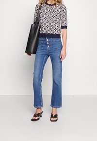 Svart tygväska, mönstrad kortärmad stickad topp, blå jeans med hög midja och svarta sandaletter med klack. Material och färg tydligt framhävt.