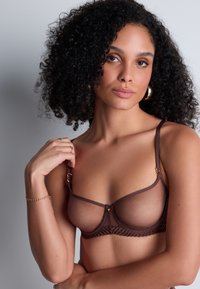 Soutien-gorge marron transparent avec des détails en dentelle délicate, des bretelles ajustables et des accents en métal doré. Texture lisse avec un design minimaliste.