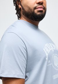 Άνδρας με γένια και dreadlocks, που φοράει γαλάζιο t-shirt με λευκή επιγραφή "Jack & Jones" και σχέδιο κοχυλιού στο στήθος.