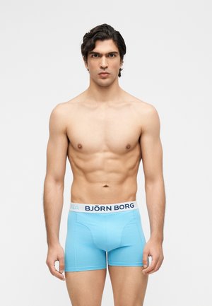 Umięśniony mężczyzna o ciemnych włosach, ubrany w jasnoniebieskie bokserki Björn Borg, stojący na tle jednolitego białego tła, zwrócony przodem.