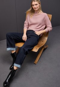 Blondynka w różowym prążkowanym swetrze, ciemnych jeansach z podwiniętymi nogawkami i czarnych butach, siedząca w zrelaksowanej pozycji na drewnianym krześle na tle w ciemnym szarym kolorze.