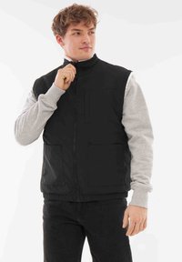 Svart vadderad väst med hög krage, dragkedjestängning och två framfickor; bärs över en grå långärmad tröja. Slätt, lätt tyg.