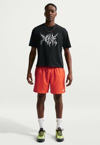 Νέος άνδρας όρθιος, φοράει μαύρο μπλουζάκι με γραφικά, φωτεινά πορτοκαλί σορτς Nike, λευκές κάλτσες Nike και νέον πράσινα αθλητικά παπούτσια σε απλό φόντο.