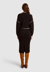 Czarny zestaw swetra dzianinowego i midi spódnicy z kontrastującymi jasnymi akcentami, cechujący się dopasowanym krojem i rozcięciem z tyłu. Modelka nosi beżowe kozaki sięgające za kolano.