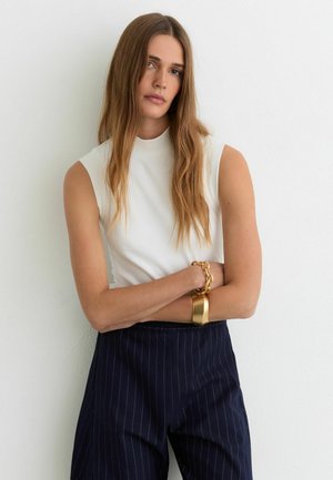 Femeie cu păr lung șaten deschis, purtând un top alb fără mâneci, pantaloni bleumarin cu dungi fine și o brățară cu lanț auriu, stând cu brațele încrucișate.