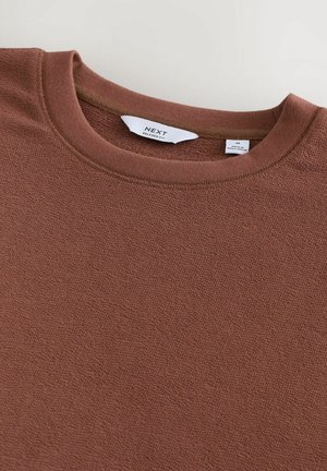 Brauner, strukturierter Pullover mit Rundhalsausschnitt. Das Etikett zeigt "NEXT" und Größe M. Der Stoff wirkt weich und leicht dehnbar.