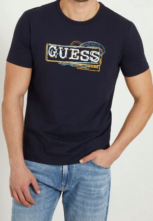 Homme portant un t-shirt Guess bleu marine avec un imprimé logo vintage multicolore et un jean bleu clair, main droite dans la poche, bras gauche détendu.