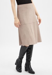 Femme portant une jupe midi beige et des bottes en cuir noir à talons hauts, se tenant devant un fond blanc.