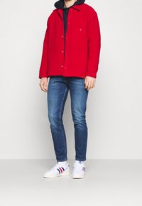 Röd knapprad jacka med två framfickor, bärs över en marinblå hoodie. Kombineras med blåjeans och vita sneakers med röda och blå ränder.