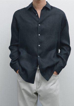 Personne portant une chemise noire à manches longues boutonnée, légèrement rentrée dans un pantalon gris clair, les mains dans les poches.