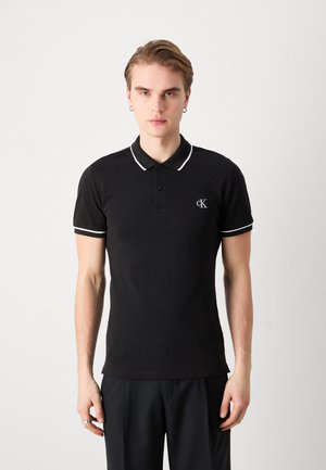 Jaunas vyras vilki juodą Calvin Klein polo marškinėlį su baltais apvadais ant apykaklės ir rankovių, stovi priešais šviesią vientisą foną.