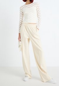 Bílý krajkový top s dlouhým rukávem se vzorem cikcak, kombinovaný s volnými krémovými kalhotami s širokými nohavicemi a bílými teniskami. Drží malou kabelku.