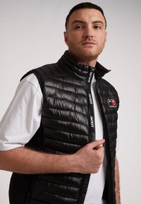 Gilet nero imbottito con colletto alto, chiusura con zip e dettagli con logo. Presenta una texture trapuntata e una maglietta bianca a contrasto.