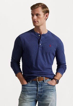 Mørkeblå langermet henley-skjorte med rund hals, med to knapper og en liten rød logo. Kombinert med falmede blå jeans og et brunt belte.