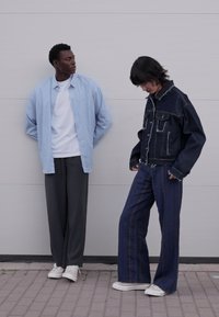 To modeller står side om side. Den ene har på seg en lyseblå stripet overskjorte og grå bukser. Den andre har på seg en mørk denimjakke og vide jeans.