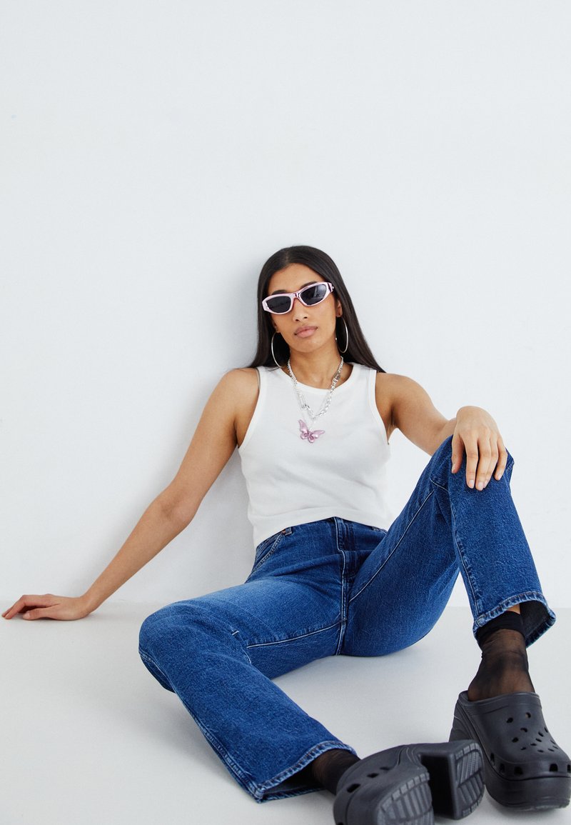 Witte mouwloze top, blauwe spijkerbroek, roze zonnebril, vlinderketting, zwarte platformklompen, tegen een eenvoudige witte achtergrond.