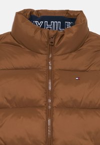 Brun puffjacka med hög krage, framdragkedja och liten logotypdetalj. Slät, glänsande tyg med quiltad struktur.
