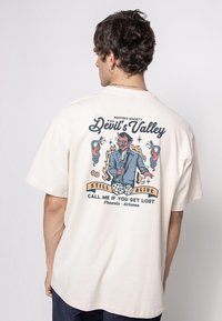 Moški nosi krema T-shirt s grafiko hudiča v obleki, ki drži mikrofon, obdanega z ognjem, kačami, kockami in besedilom.