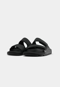 Et par svarte sandaler med to justerbare stropper og tykk såle, vinkelrett for å vise front- og sidedetaljer mot en ensfarget bakgrunn.