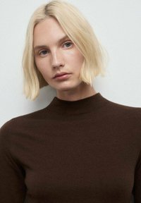 Maglione a collo alto a coste marrone, design aderente, texture morbida, modello con capelli biondi fino alle spalle, sfondo neutro.