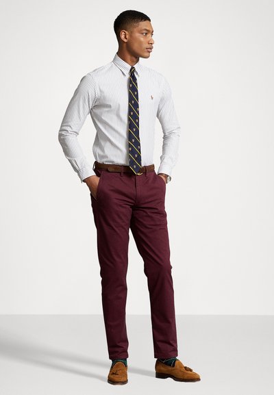 Îmbrăcăminte masculină care include o cămașă albă cu dungi albastre, o cravată bleumarin cu accente galbene, pantaloni burdundii, pantofi maro și o curea maro.