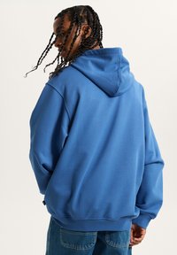 Sudadera azul hecha de un tejido suave y grueso, con capucha ajustable, puños acanalados y un corte relajado. Llevada con pantalones de mezclilla.