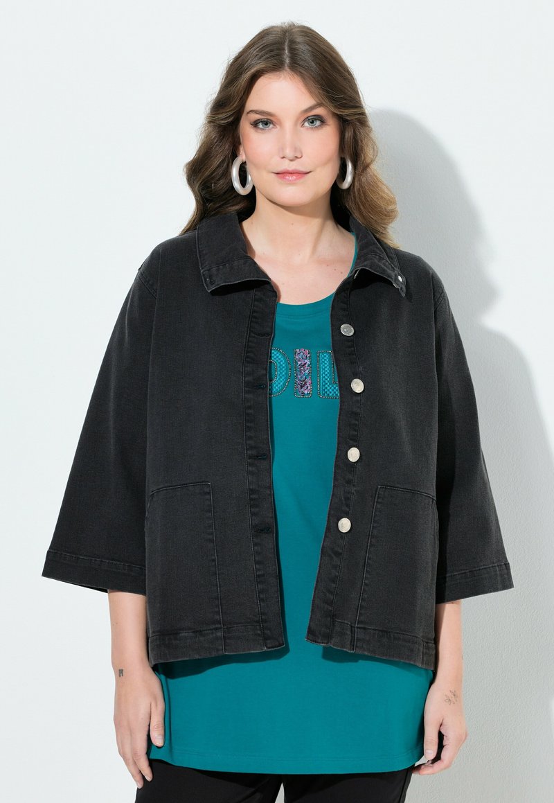 Žena s dlouhými vlnitými vlasy, která nosí stříbrné kruhové náušnice, černou sako s knoflíky a tříčtvrtečními rukávy a tyrkysovou košili s dekorativním textem.