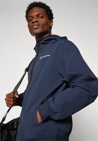 Navy blue windbreaker jacket with a zip front, hood, and elastic cuffs. Features a Champion logo in white on the chest and side pockets.