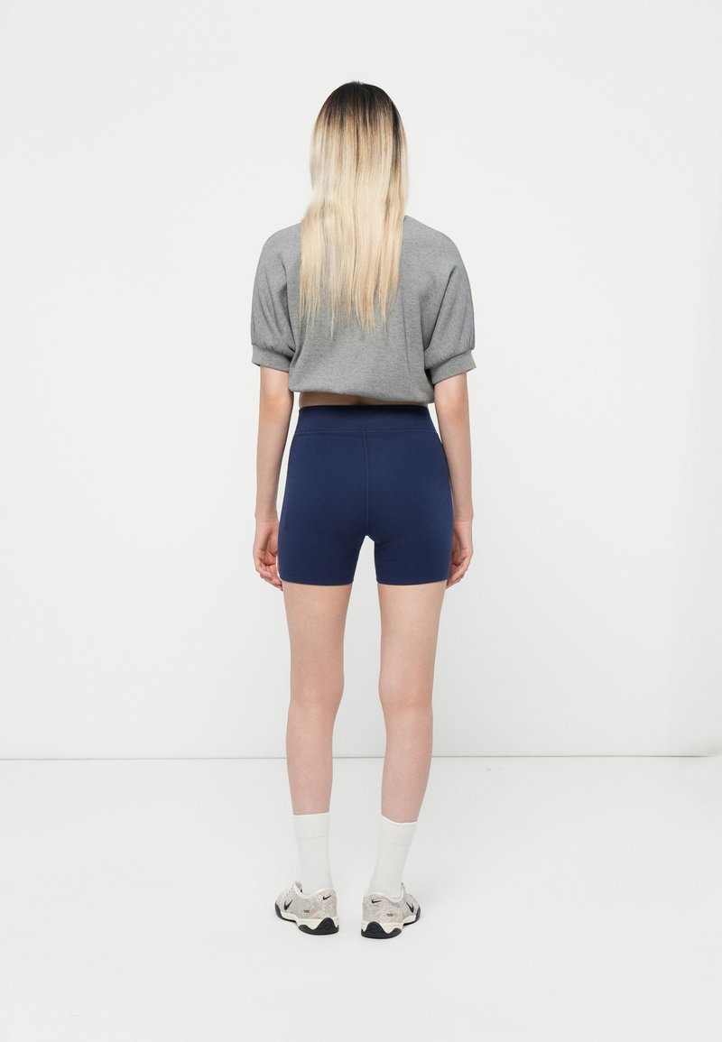 Haut gris à manches courtes et short taille haute bleu marine. Tissu lisse avec une silhouette ajustée. Fond blanc et chaussures claires avec des chaussettes.