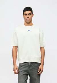 Kremfarget kortermet sweatshirt med ribbestrikket hals og kant, med en liten blå logo i midten. Kombinert med grå jeans.