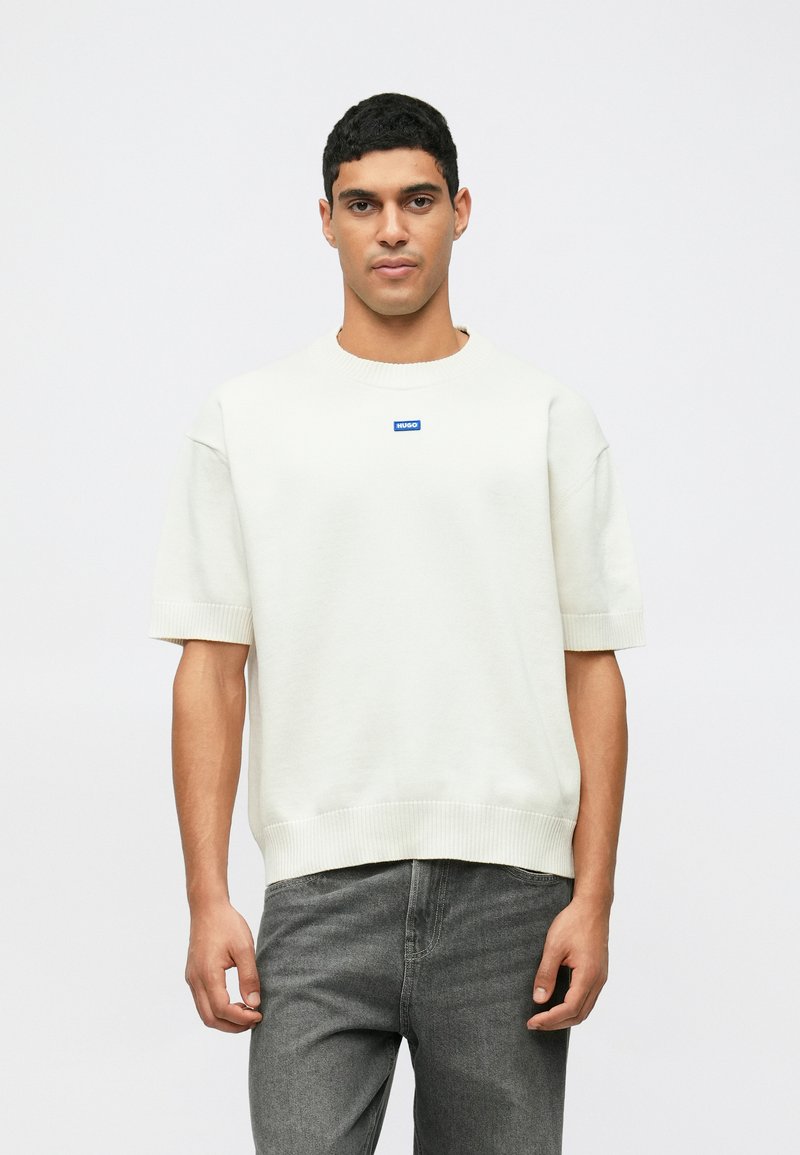 Kremfarget kortermet sweatshirt med ribbestrikket hals og kant, med en liten blå logo i midten. Kombinert med grå jeans.