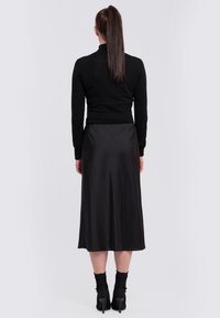 Haut noir à manches longues associé à une jupe midi en satin noir fluide, présentant une texture lisse et des bottes à talons noires arrivant à la cheville.