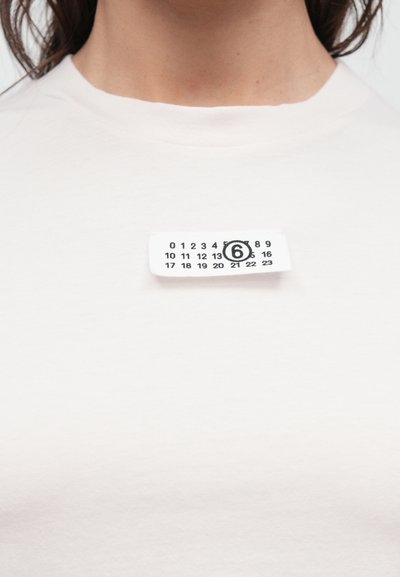 Vaaleabeige pyöreäkauluksinen t-paita. Etupuolella on pieni valkoinen etiketti, jossa on numeroviiva, musta ympyrä ja numero "6".