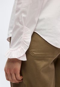 Main reposant le long du corps, portant un chemisier rose clair avec un poignet à volants et un pantalon marron avec une couture visible sur la poche arrière.